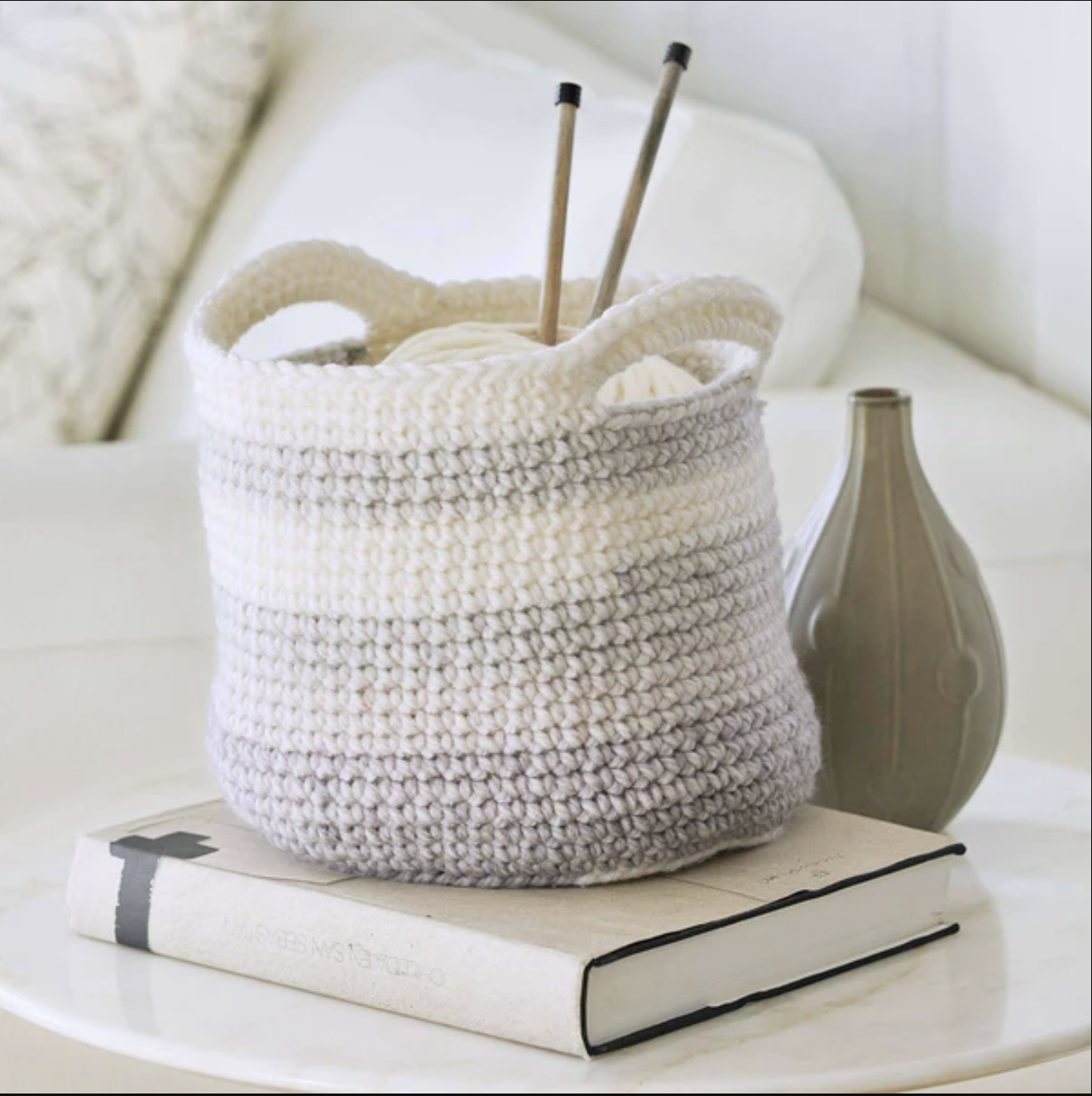 A beginner crochet basket with handles, holding two knitting needles and cream-colored yarn, sits atop a closed book. Beside the basket is a decorative vase. The items rest on a white surface in a softly lit room. -Marly Bird